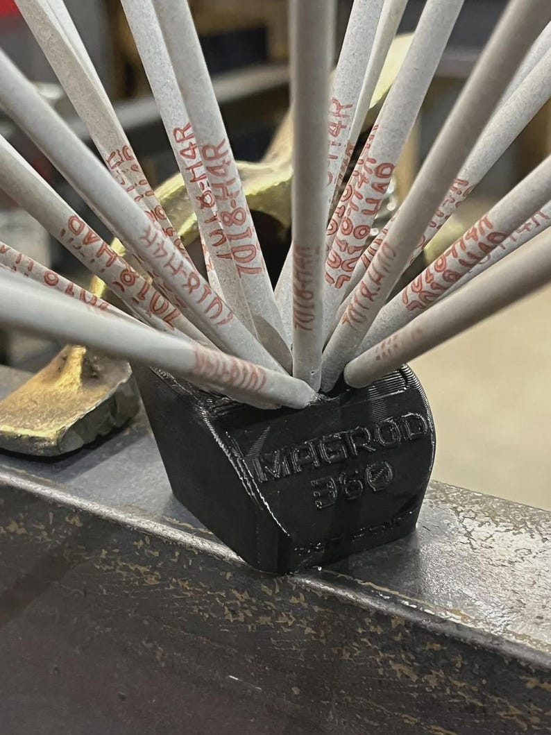 May include: A black MAGROD 360 holder containing multiple welding rods. The rods are light gray with red text. The holder is on a metal surface.