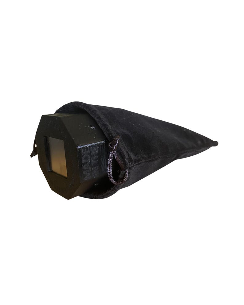 May include: A black, geometric prism with a rectangular window, partially enclosed in a black velvet drawstring bag. The text "MADE IN THE" is visible on the prism. The bag is closed with a black drawstring.