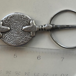 May include: A silver-toned metal brooch featuring a central circular coin-like element with engraved Arabic script and the number 1299, attached to a pin and a circular ring. A ruler shows the object measures approximately 4.5 inches in total length.