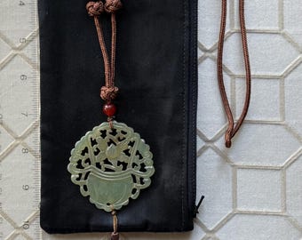 Op de afbeelding: Een jade hanger ketting met een gebeeldhouwd bloemenontwerp, bruin koord en kwast. De ketting heeft houten en rode kralen accenten. De hanger wordt getoond op een zwarte pouch.