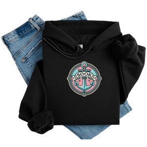 Puede incluir: Sudadera con capucha negra con un gráfico de un ancla azul con un círculo de cuerda rosa y blanco alrededor. El texto "SOOK" está en una pancarta blanca.