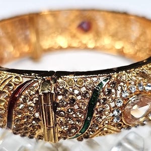 May include: Close-up of a gold-colored hinged bangle bracelet. The bracelet features intricate filigree and is adorned with clear and amber-colored gemstones, as well as red and green enamel accents. The clasp is visible.