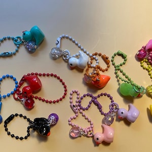 May include: A collection of colorful duck-shaped keychains in various colors, including blue, white, orange, green, pink, purple, and yellow. Each keychain features a beaded chain and decorative accents, such as clear beads and glitter.