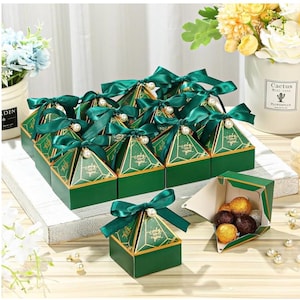 May include: Green and gold pyramid-shaped favor boxes with green satin ribbon and pearl accents. The boxes are filled with chocolate candies.