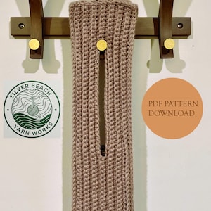 May include: A light brown, crocheted item with a vertical slit, hanging on a wooden coat rack with brass knobs. The item is displayed against a white wall. A circular logo and a text that says "PDF PATTERN DOWNLOAD" are also visible.