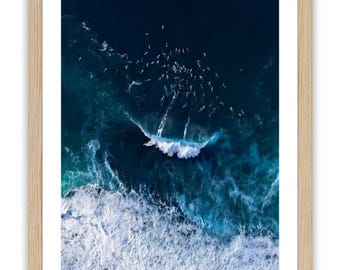 Può includere: Fotografia aerea di surfisti nell'oceano, incorniciata in legno chiaro. L'immagine cattura un gruppo di surfisti che cavalcano le onde nel mare blu scuro. Le onde bianche e schiumose si infrangono vicino alla riva. L'immagine è firmata in basso a destra.