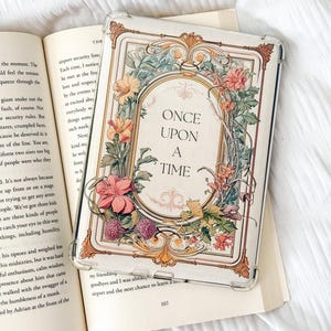 Può includere: Custodia trasparente per telefono con un design floreale e la scritta "Once Upon a Time" in una cornice ovale. La custodia è sopra un libro aperto. La custodia ha un bordo decorativo con fiori.