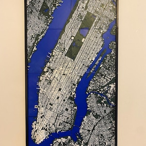 Può includere: Una mappa tridimensionale incorniciata di New York City. La mappa presenta una rappresentazione dettagliata della disposizione a griglia della città, con edifici bianchi e parchi verdi su uno sfondo d'acqua blu. La mappa è racchiusa in una cornice nera.