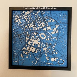 May include: A framed 3D map of the University of North Carolina, featuring a blue background and white building outlines. The black frame surrounds the detailed campus layout, including roads and landmarks. The text "University of North Carolina" is at the top.