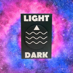 Puede incluir: Un letrero rectangular negro con las palabras "LIGHT" y "DARK" en blanco, en negrita y mayúsculas. Un círculo negro con un triángulo blanco y tres líneas onduladas está en el centro. El fondo es una galaxia colorida.