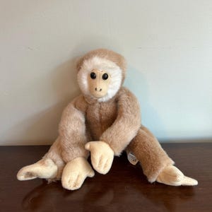 May include: A plush monkey toy with a light brown body and cream-colored face, hands, and feet. The stuffed animal has black eyes and is sitting on a dark brown surface.