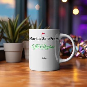 Puede incluir: Taza de cerámica blanca con el texto "Marked Safe From The Rapture" en negro y verde, con una bandera roja. La palabra "Today" está impresa debajo. La taza está sobre una superficie de madera.
