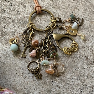 May include: A gold-toned charm bracelet with a variety of charms, including keys, hearts, leaves, and stones. The bracelet is attached to a brown leather cord.