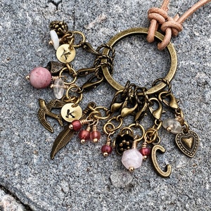 May include: A gold-toned charm necklace with a leather cord. The necklace features a variety of charms, including a heart, a feather, a bird, and initial charms. The charms are attached to a large, round ring.
