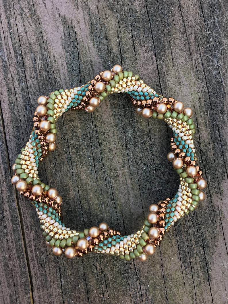 K&ouml;nnte beinhalten: Ein Armband mit Perlen in Spiralform. Das Armband besteht aus kleinen, goldenen, gr&uuml;nen und blauen Perlen. Das Armband ist mit gr&ouml;&szlig;eren, kupferfarbenen Perlen verziert.
