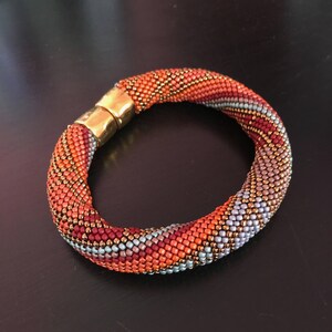 May include: A colorful beaded bracelet with a gold clasp. The bracelet is made with small, round beads in shades of orange, red, blue, and brown. The beads are arranged in a spiral pattern.