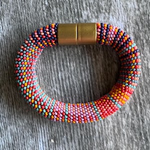 May include: A tubular beaded bracelet featuring a vibrant, multi-coloured pattern of small glass beads in shades of coral, pink, orange, light blue, and dark purple. The bracelet is secured with a cylindrical, matte gold-toned metal magnetic clasp.