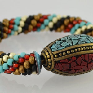 May include: A multi-colored beaded bracelet with a large, oval-shaped bead in the center. The bead is made of red, blue, and turquoise mosaic tiles and has a gold-colored rim.