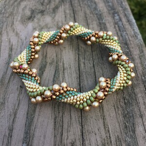 K&ouml;nnte beinhalten: Ein Armband mit einem Spiraldesign in Gr&uuml;n-, Gold- und Braunt&ouml;nen. Das Armband ist aus kleinen Perlen und gr&ouml;&szlig;eren Perlen gefertigt, die an Perlen erinnern.