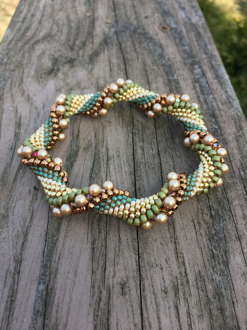 K&ouml;nnte beinhalten: Ein Armband mit einem Spiraldesign in Gr&uuml;n-, Gold- und Braunt&ouml;nen. Das Armband ist aus kleinen Perlen und gr&ouml;&szlig;eren Perlen gefertigt, die an Perlen erinnern.