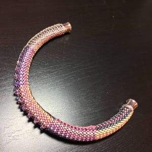 Peut inclure: Un collier multi-colore en perles avec un dégradé de rose, jaune et noir. Le collier est composé de petites perles carrées et possède un fermoir argenté.