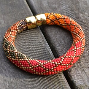May include: A beaded bracelet with a geometric pattern in shades of red, orange, gold, and green. The bracelet has a gold clasp.