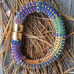 Puede incluir: Un brazalete de cuentas de colores con un cierre dorado. El brazalete está hecho de pequeñas cuentas redondas en tonos de morado, azul, verde, amarillo y naranja. Las cuentas están dispuestas en espiral.