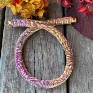 May include: A beaded necklace with a gradient of purple and gold beads. The necklace is made of small, round beads and has a gold clasp.