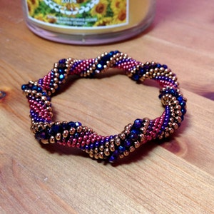 May include: A close-up of a beaded bracelet with a spiral design. The bracelet is made with blue, pink, and gold beads.