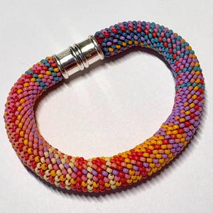 May include: A tubular beaded bracelet featuring a vibrant, multi-coloured pattern of small glass beads in shades of pink, orange, yellow, purple, and blue. The bracelet is secured with a polished silver-toned cylindrical magnetic clasp.