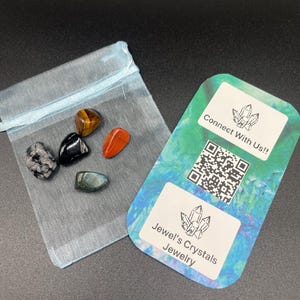 Peut inclure: Un assortiment de pierres précieuses polies dans un sac en filet bleu clair, à côté d'une carte de visite. Les pierres précieuses comprennent l'œil de tigre, l'obsidienne, la cornaline et l'obsidienne flocon de neige. La carte de visite porte le texte "Connectez-vous avec nous !!" et "Jewel's Crystals Jewelry".