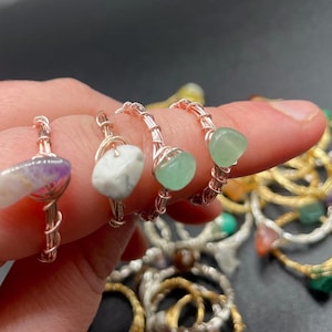 May include: Handmade wire-wrapped rings with natural gemstones. The rings feature amethyst, white, and green stones. The wire is a copper colour. Other rings with various gemstones are in the background.