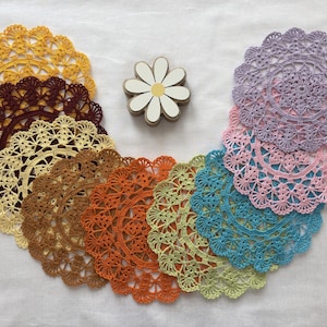 May include: A collection of colorful, handmade crochet doilies in shades of yellow, brown, orange, green, blue, pink, and purple. A wooden daisy-shaped accent is placed among the doilies. The doilies are arranged on a white surface.