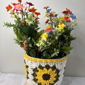 May include: A colorful floral arrangement in a crocheted pot. The pot features a sunflower design with yellow, white, and green details. The flowers are a mix of red, orange, yellow, pink, and blue, creating a vibrant display.