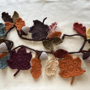 May include: A crocheted garland featuring autumn leaves and acorns in various colors. The leaves are in shades of brown, orange, yellow, and green, with gray acorns. The garland is a decorative item for fall or Thanksgiving.
