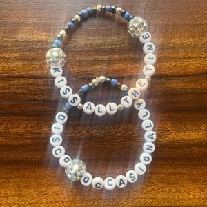 May include: Two beaded bracelets. One bracelet spells out "DISCO OCCASIONS" and the other spells out "KISS ALL THE TIME" in white letter beads. Both bracelets have disco ball beads and blue and silver accent beads.