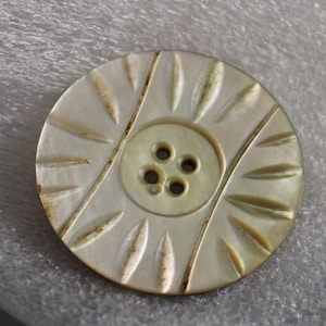 May include: A round, off-white button with a central button and four holes. The button has a decorative pattern of diagonal lines and leaf-shaped designs radiating from the center. The button measures approximately 1 inch in diameter.