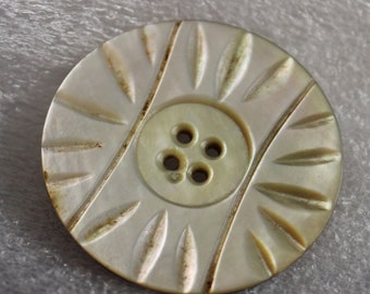 May include: A round, off-white button with a central button and four holes. The button has a decorative pattern of diagonal lines and leaf-shaped designs radiating from the center. The button measures approximately 1 inch in diameter.