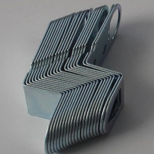 Puede incluir: Una pila de soportes metálicos plateados en forma de L. Cada soporte tiene una curva en ángulo recto y un agujero circular en un extremo. Los soportes están cuidadosamente dispuestos, mostrando sus bordes y los agujeros.