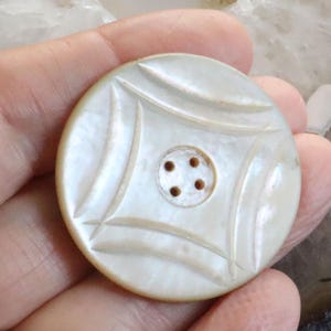 May include: A round, off-white button with a carved design. The button features a central circle with four holes and an outer pattern of curved lines and diamond shapes. The button is held in a hand.