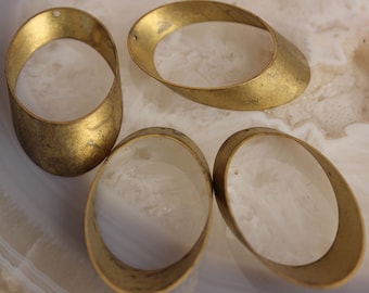 May include: Four oval-shaped, gold-colored metal jewelry findings. Each finding has a curved, open design with a textured surface. Two findings have a small hole at the top. The findings are arranged on a white and gray surface.