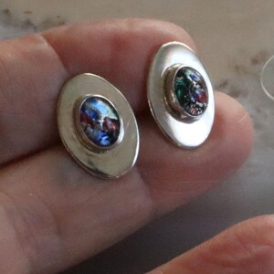 May include: A pair of oval-shaped stud earrings. Each earring features a colorful, speckled, oval-shaped center stone set within a silver-toned bezel. The earrings have a polished, metallic finish, reflecting light.