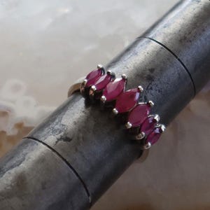 May include: A silver ring featuring a series of marquise-cut, deep red gemstones. The gemstones are set in a row, creating a continuous pattern. The ring is displayed on a dark, metallic surface, highlighting the contrast between the bright gemstones and the metal.