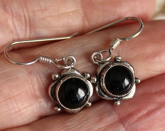 May include: A pair of silver earrings with black, round stones. The earrings have a decorative, floral-like design around the stones and a simple hook closure. The earrings are shown against a neutral background.