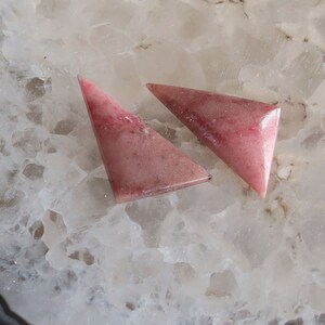 May include: Two triangular, polished gemstones in shades of pink and red. The stones have a smooth, glossy surface and a marbled appearance. The gemstones are set against a white, textured background.