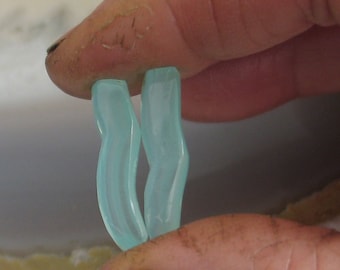 May include: Two light blue, curved glass pieces are held between fingers. The pieces have a smooth, glossy finish and a slight wave shape. The background is out of focus, highlighting the glass.