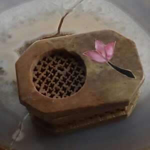 May include: A small, octagonal, brown stone box with a carved, circular opening and a pink and black floral inlay. The box is made of a light brown stone and has a decorative, carved design on the top.