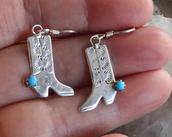 May include: A pair of silver-toned cowboy boot earrings. Each earring features a detailed boot design with a turquoise-colored stone accent. The earrings are held by a person's hand.