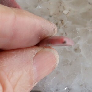 May include: A small, pink, polished gemstone, held between fingers. The stone is a light pink color with a darker pink area and a small black inclusion. The gemstone is oval-shaped and appears to be translucent.