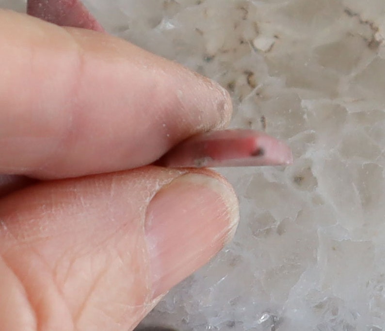 May include: A small, pink, polished gemstone, held between fingers. The stone is a light pink color with a darker pink area and a small black inclusion. The gemstone is oval-shaped and appears to be translucent.